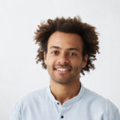 Headshot of good-looking positive young dark-skinned male with stubble and trendy haircut wearing blue shirt while posing isolated against blank studio wall background with copy space for your text